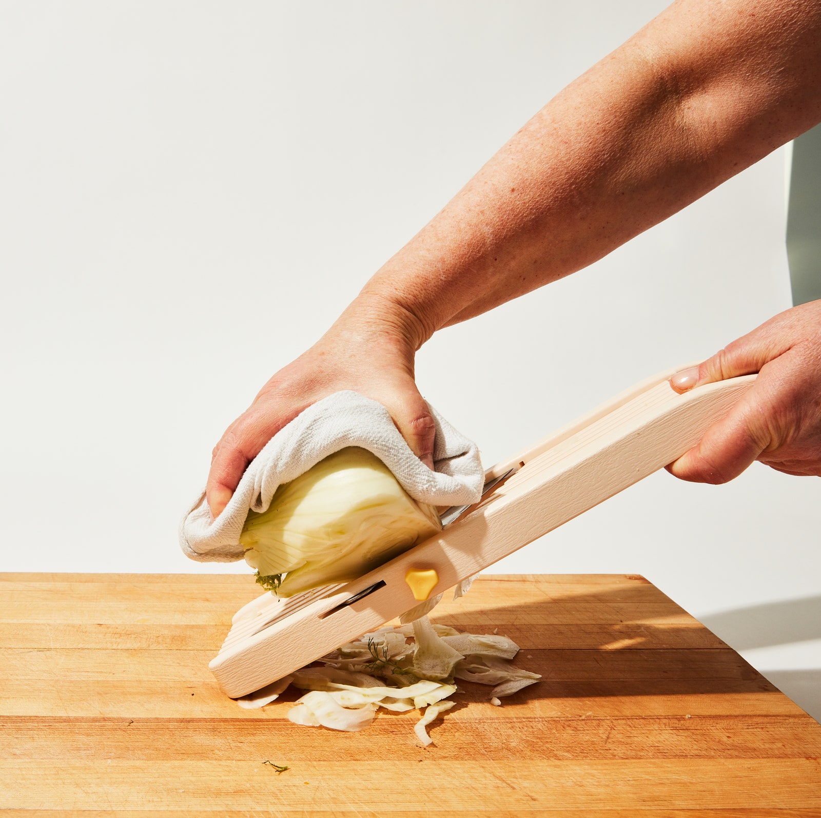 A hand shaving a half bulb of fennel on a mandoline using a kitchen towel as a protective guard.